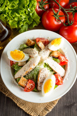 Fresh salad made of tomato, ruccola, chicken breast, eggs, arugula, crackers and spices. Caesar salad in a white, transparent bowl on wooden background