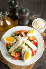 Fresh salad made of tomato, ruccola, chicken breast, eggs, arugula, crackers and spices. Caesar salad in a white, transparent bowl on wooden background