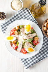Fresh salad made of tomato, ruccola, chicken breast, eggs, arugula, crackers and spices. Caesar salad in a white, transparent bowl on wooden background