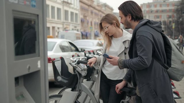 Happy romantic couple of tourists taking bikes for rent in city street, use phone application and credit card, man and woman rent bycicles