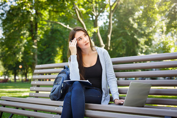 Young female IT student on a bench,. Doing homework in the park with laptop and textbook