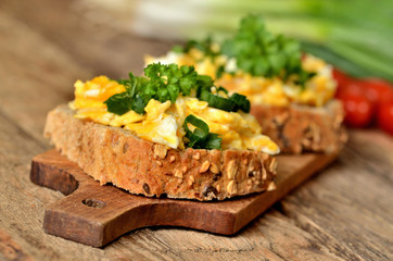 Close-up of wholemeal slices of bread with scrambled eggs, fresh herbs, spring onions and tomatoes in background