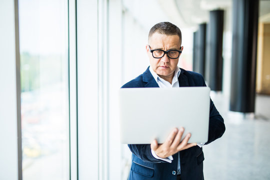 Always ready to help you. Cheerful mature man in formalwear adjusting his eyeglasses and holding laptop while standing in office