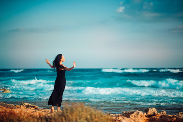 Lonely inspired woman in black dress staying on seaside dreaming and looking to sea on windy sunset day. Back view. Emotions loneliness Lifestyle Solitariness concepts