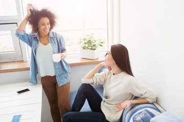 Two students are talking to each other. Afro american girl is standing near window and touching her hair while european girl issitting and looking at her friend.