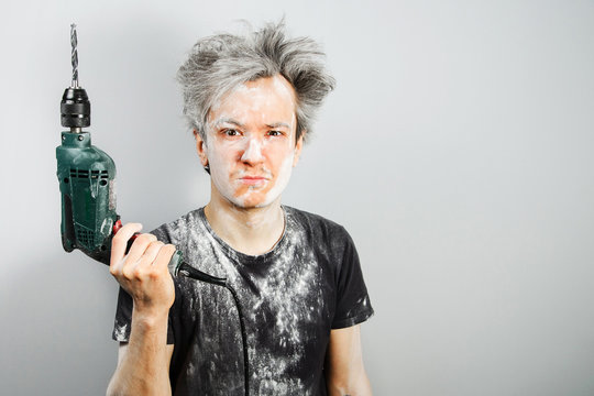 Unshorn and unshaven young guy soiled in plaster, holded drill and on gray background.
