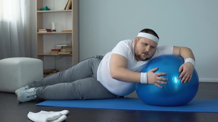 Tired unmotivated obese man lying on fitness ball, slimming fitness program