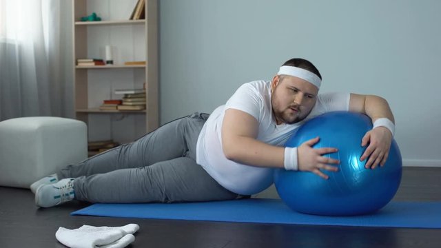 Tired unmotivated obese man lying on fitness ball, slimming fitness program