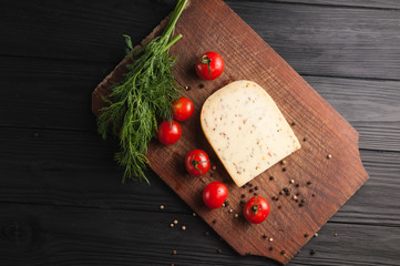 Cheese on a brown wooden board on a black wooden background, cherry tomatoes, pepper, greens, spices, dill, parsley. View from above.