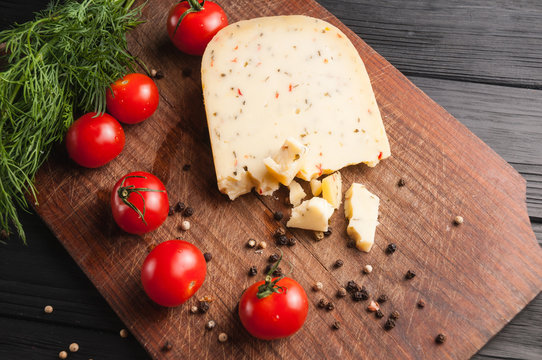 Cheese on a brown wooden board on a black wooden background, cherry tomatoes, pepper, greens, spices, dill, parsley. View from above.