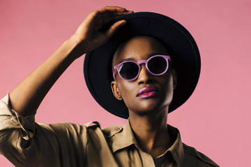 A fashion portrait of a young woman with purple sunglasses and black hat, isolated on pink studio background  - Powered by Adobe
