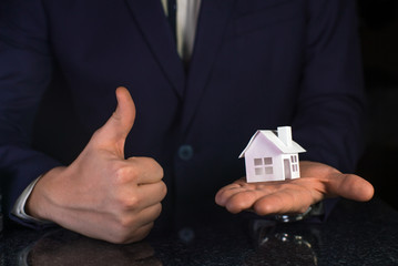 Businessman shows thumb up with small paper house in his hand. Good real estate concept