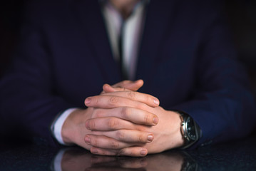 Obraz premium Businessman in blue suit and white shirt with crossed fingers sitting by the table on dark background