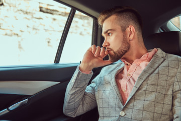 Successful stylish young businessman in a gray suit and pink shirt, riding on a back seat in a luxury car.