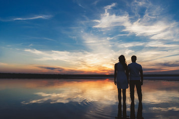 Portrait, silhouette of happy couple watching the colourful bright sunset standing in large lake, reflection in the water, holding hands - Powered by Adobe
