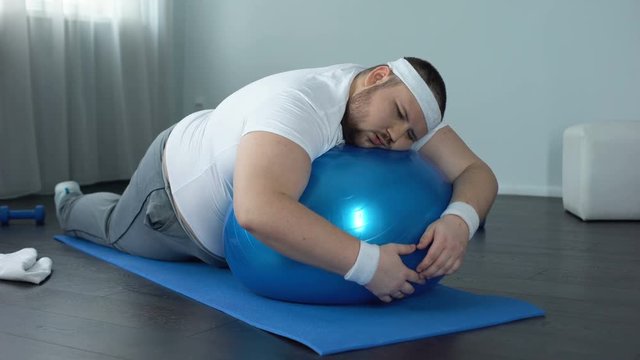 Weak-willed fat man relaxing on fitness ball, home workout break, laziness