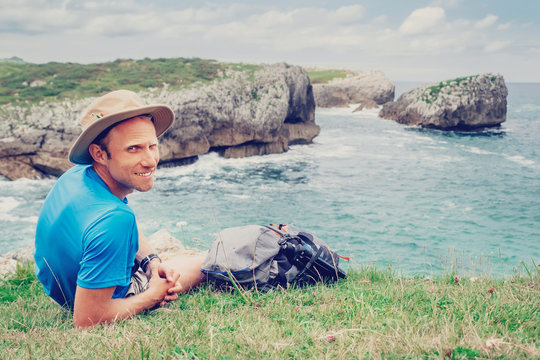Man backpacker traveler rests on the rocky sea side, looks in camera and smile