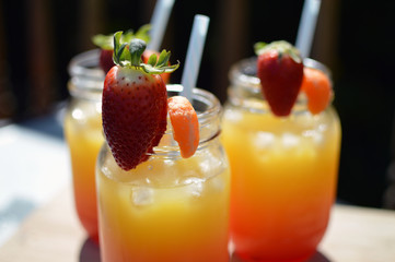 Three summer alcoholic cocktails on a wooden table in the sunlight. Orange and red in colour with fresh strawberry and orange segment on top of glass. Straw in beaker. 