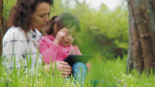 Family in nature with a tablet. Mom with a little daughter on green grass with a tablet.