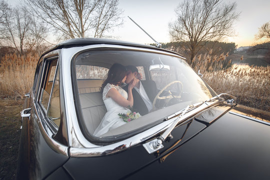 Young wedding couple sitting smiling inside retro car and are kissing each other. just married embrace is hugging inside car. bride hugging groom who is driving the car. Wedding with retro car.