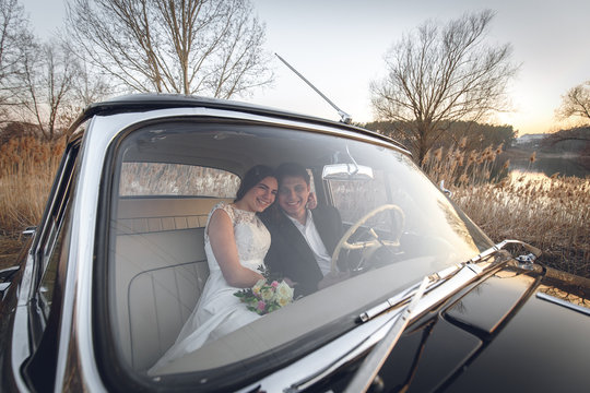 Young wedding couple sitting smiling inside retro car and looking at each other. just married embrace is hugging inside car. bride hugging groom who is driving the car. Wedding with retro car.