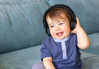 Adorable little baby boy listening to music in headphones on his head sitting on blue sofa at home and smiling with his first eight teeth, musical education concept,