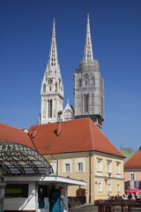 Centrum Zagrzebia, stolicy Chorwacji, z budynkiem zadaszonym pasażem oraz widocznymi zza budynku dwiema wieżami katedry katolickiej, jedna z wież restaurowana, z rusztowaniem, błękitne niebo - obrazy, fototapety, plakaty