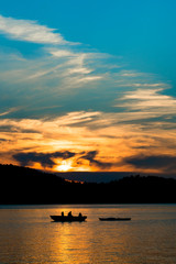 Sunsets through the clouds, while dad fishes with children (in silhouette) from a boat on the lake, while towing a kayak.