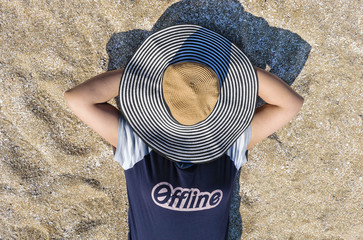 Woman offline and disconnected on the beach sand with hat covering her face - obrazy, fototapety, plakaty