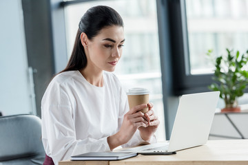 Fototapeta premium young businesswoman holding paper cup and using laptop at workplace