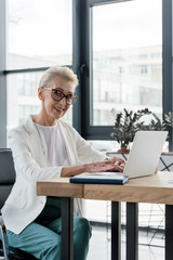 Fototapeta na wymiar cheerful senior businesswoman in eyeglasses using laptop and smiling at camera in office