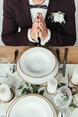cropped shot of groom in suit praying while sitting at served table, rustic wedding concept