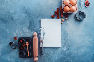 Pastry recipe concept with baking tools, ingredients and a notepad with blank pages on a modern background with copy space. Scoops, whisks, cookie cutters, muffin tin, eggs and cinnamon from above.