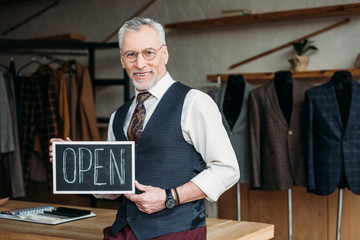 handsome mature tailor holding chalkboard with open sign at sewing workshop