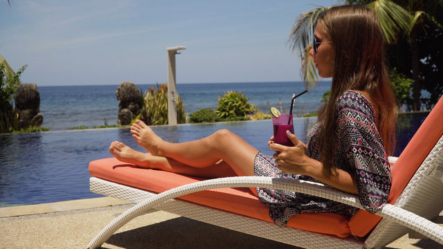 Beautiful young girl with glass of cocktail relaxing near swimming pool. Girl in sunglasses with cocktail in seafront near the bassin amongst the palm trees on the background of the sea. Travel