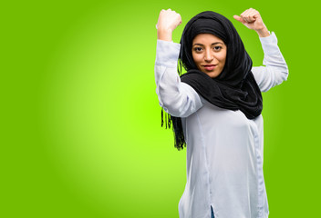 Young arab woman wearing hijab showing biceps expressing strength and gym concept, healthy life its...