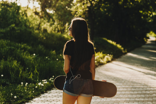 Photo from back of a beautiful young caucasian girl with a skateboard in her hands looking away at the grass and with smart phone in her back pocket.