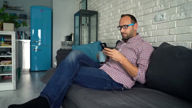 Man with smartphone drinking tea on sofa at home
