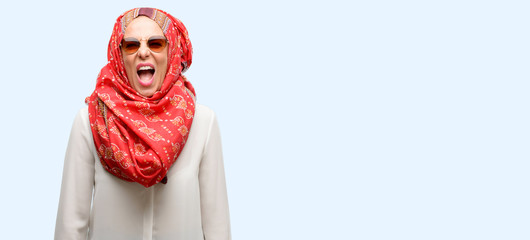 Middle age muslim arab woman wearing hijab stressful, terrified in panic, shouting exasperated and frustrated. Unpleasant gesture. Annoying work drives me crazy isolated blue background