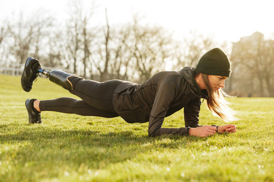 Image of sporty handicapped woman in black tracksuit, warming up and doing plank with prosthesis leg on nature