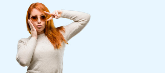 Beautiful young redhead woman looking at camera through fingers in victory gesture winking the eye and blowing a kiss isolated over blue background