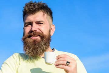 Man with long beard enjoy coffee. Coffee taster concept. Bearded man with espresso mug, drinks coffee. Man with beard and mustache on smiling face drinks coffee, blue sky background, defocused.