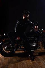 Naklejka na ściany i meble Man with beard, biker in leather jacket lean on motor bike in darkness, black background. Macho, brutal biker in leather jacket stand near motorcycle at night time, copy space. Biker culture concept.