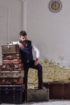 Move out and relocation concept. Man with beard and mustache packed luggage, white interior background. Macho elegant on tired face, exhausted at end of packing, leans on pile of vintage suitcases.