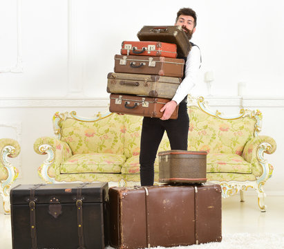 Man with beard and mustache wearing classic suit delivers luggage, luxury white interior background. Macho elegant on strict face carries pile of vintage suitcases. Butler and service concept.