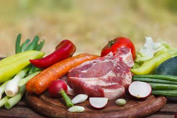 Set of fresh vegetables and raw steak meat on wooden board. Outdoor image, blurred nature in background.