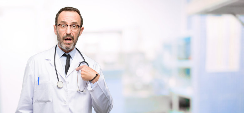 Doctor senior man, medical professional happy and surprised cheering expressing wow gesture, pointing with finger at hospital