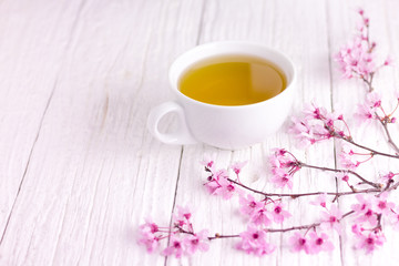 A cup of tea and blossomed cherry tree branch, using white wooden table. Slightly pink shades.