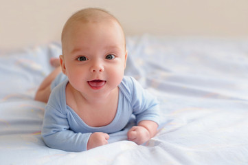 Adorable baby lying in bed in blue bodysuit and smiling cheerfully. Cute kid during tummy time. Nursery for children. Textile and bedding for kids. Family morning at home.