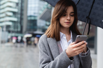 Brunette businesswoman using mobile phone near office, young girl browsing phone smiling under umbrella while it is raining outdoors, female manager texting smartphone near modern bank building